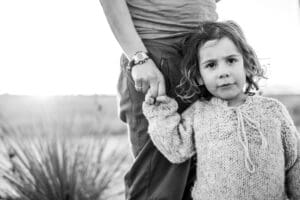 Young girl holds a parent's hand while standing outdoors at sunset, looking directly at the camera