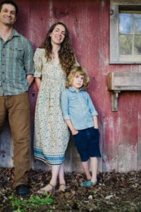 Casual family portrait with parents and child standing against a red barn wall, smiling and relaxed