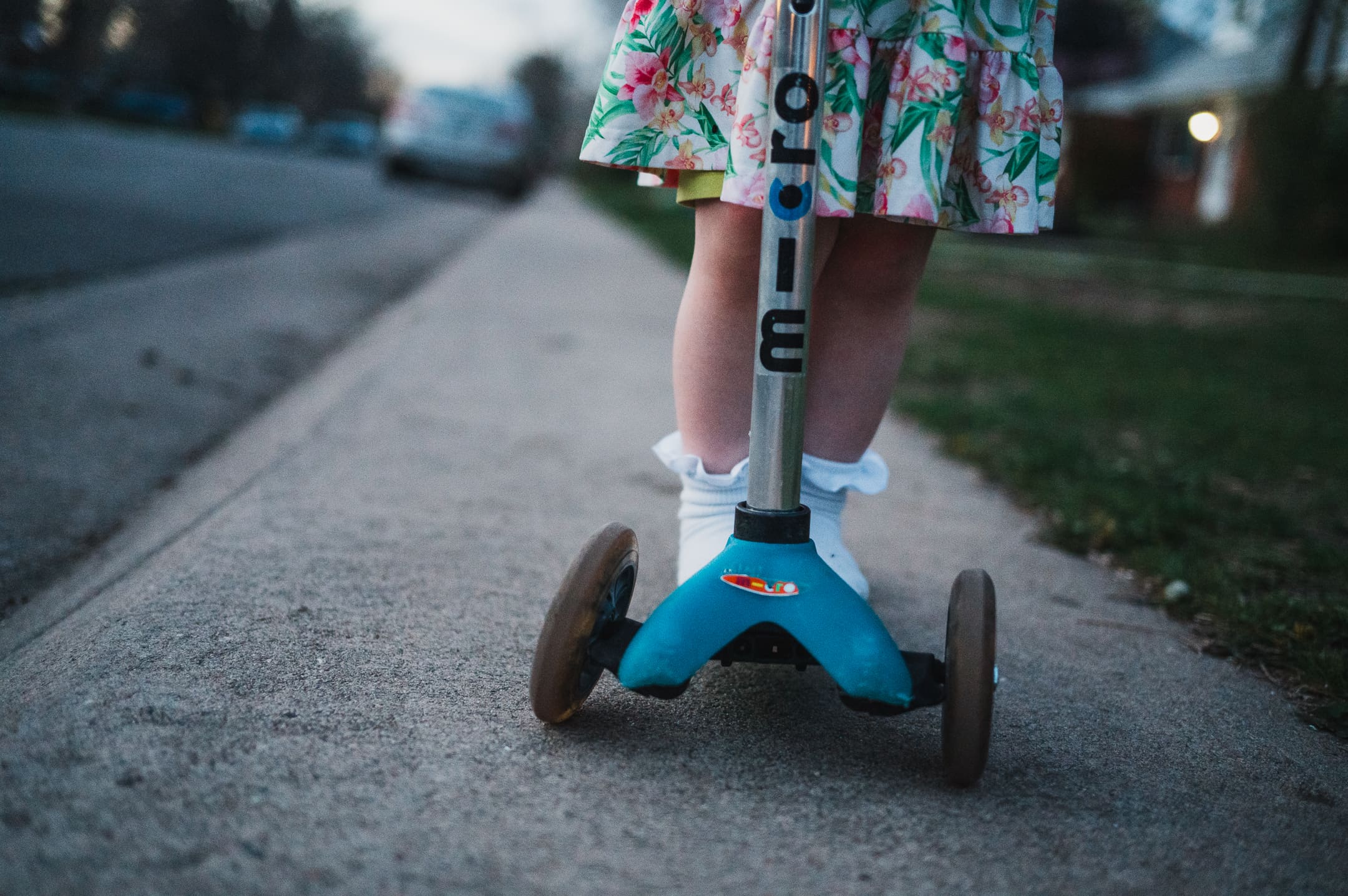A close-up of a child’s legs and scooter on a sidewalk at dusk. The child wears a floral dress and white socks. A simple, everyday detail captured in candid family photography.