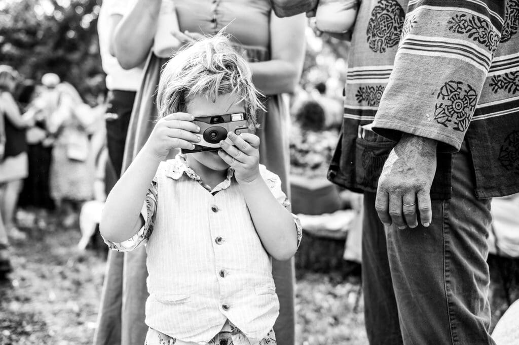 A small child holds a point-and-shoot camera up to their face, capturing a photo while surrounded by adults at a gathering. A playful example of candid family photography.