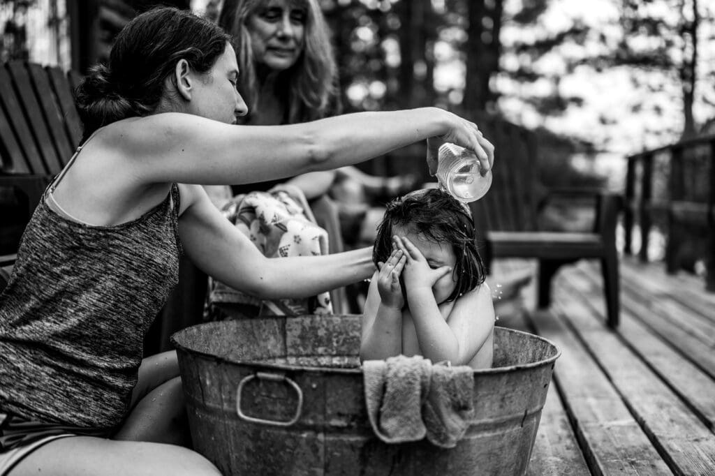 A candid moment of a child covering their face while a parent pours water over their head during an outdoor bath in a metal tub. A quiet slice of documentary family life photography.