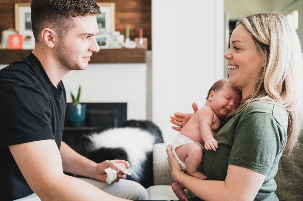 Mother smiling while holding newborn as partner sits nearby in the living room