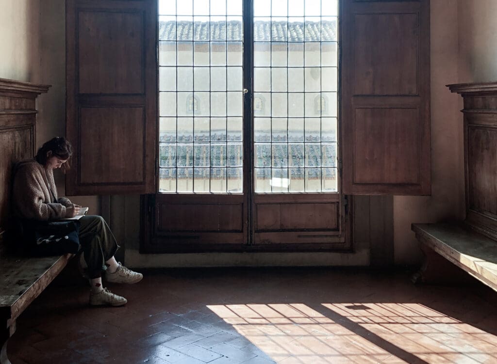 Woman writing in a sunlit room with large wooden windows in Italy