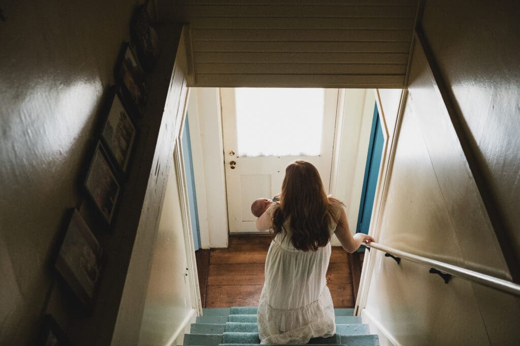 A mother in a white dress holds her newborn as she walks down a stairwell, heading toward a bright doorway. A quiet, tender moment captured in documentary newborn photography.