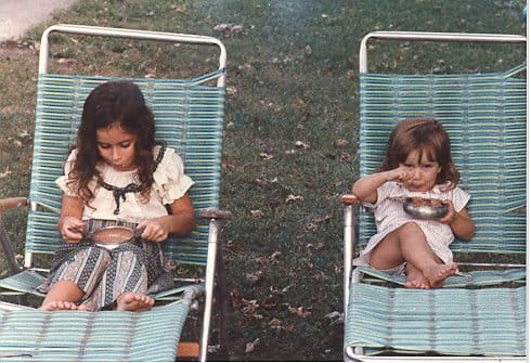 Two young sisters sit side by side in woven lawn chairs, each eating from a metal bowl on a patch of grass. A quiet, nostalgic moment from childhood.