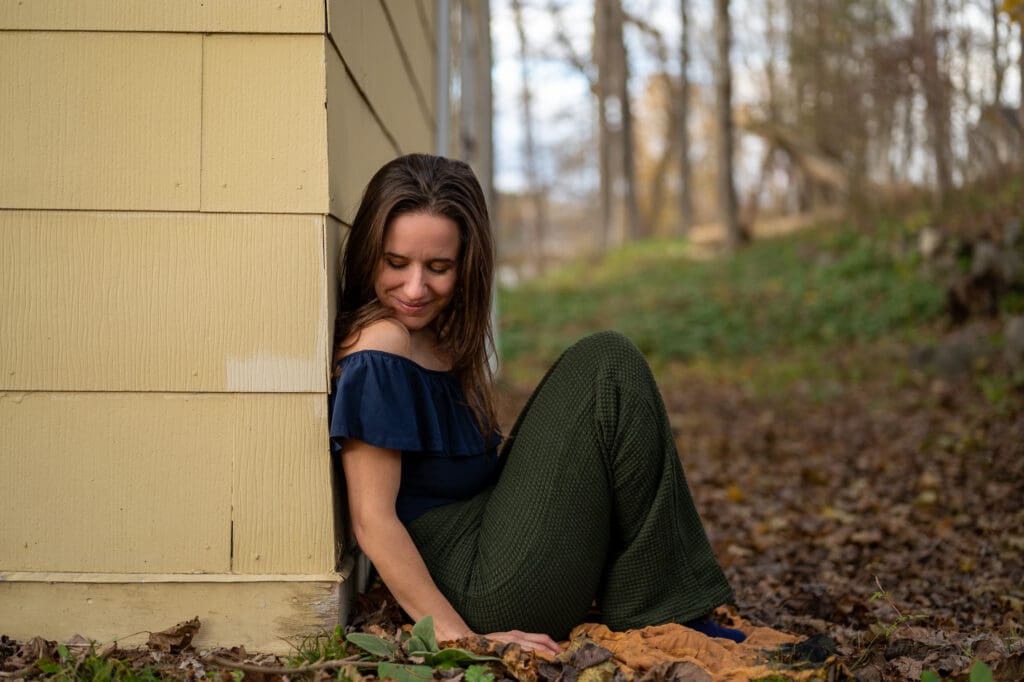 Woman in an off-the-shoulder top leaning against a yellow house wall surrounded by autumn leaves