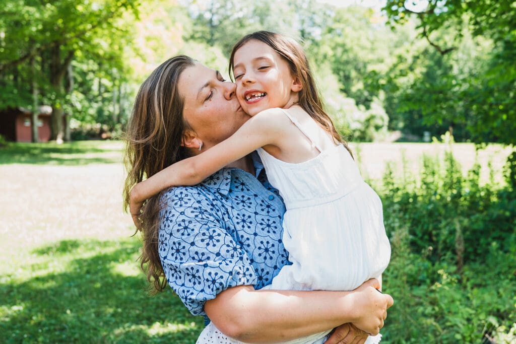 Mother hugging young daughter in white dress during outdoor family photography session