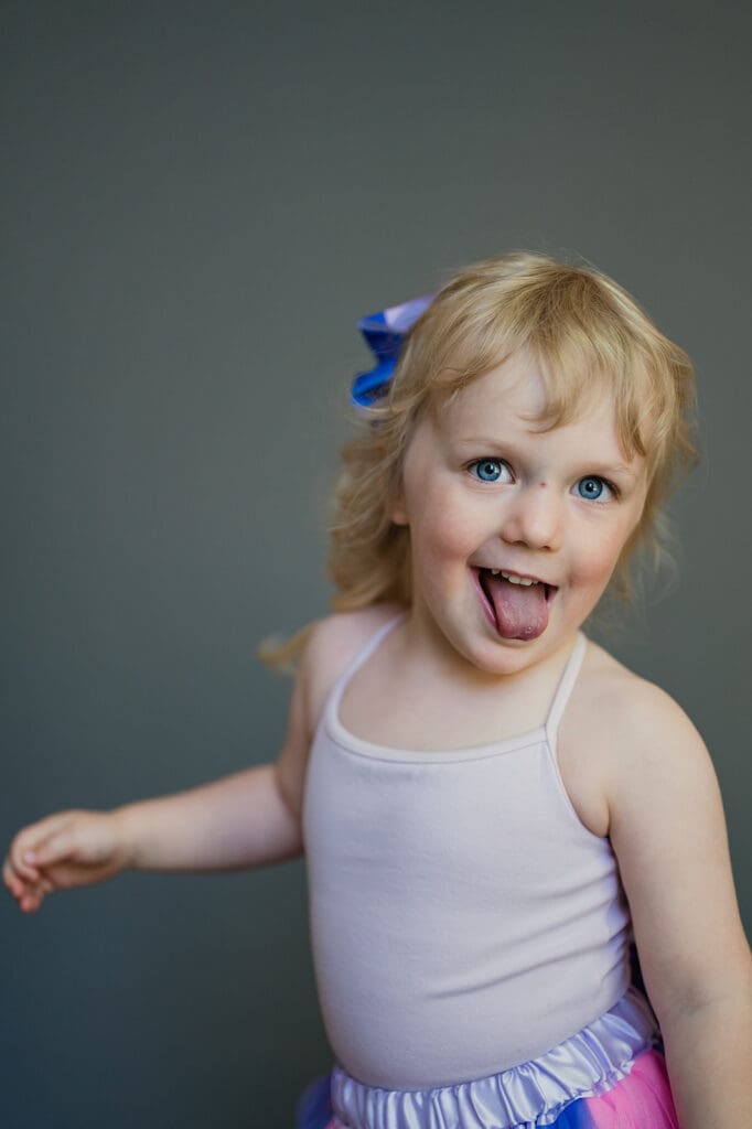 Young girl with blue eyes and bow in her hair smiling and sticking out tongue during fine art school portrait session by Rebecca Bloomfield Photography