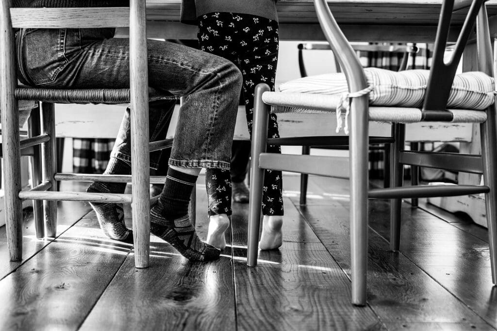 a girl on her tip toes stands next to her parent who is sitting at a table during a documentary style family photography session