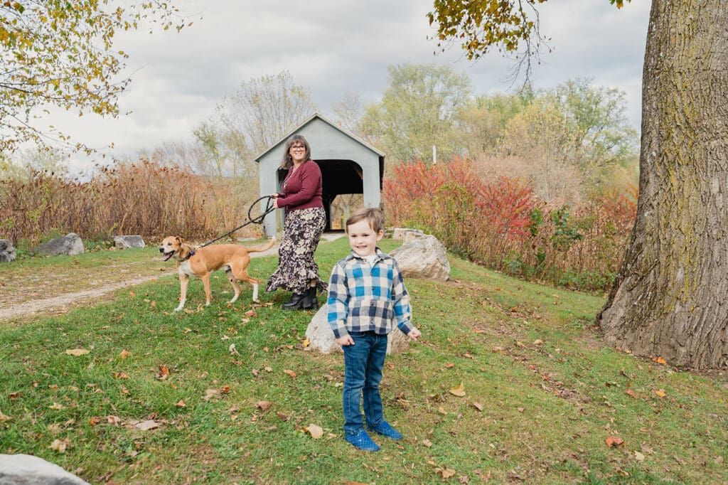 A mother walking her dog while her young child stands nearby during a lifestyle family photography session near a covered bridge