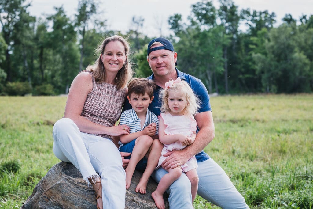a family of four sits on a large boulder outside their family home with two small kids on the parents' laps they are smiling at the camera in this family photo