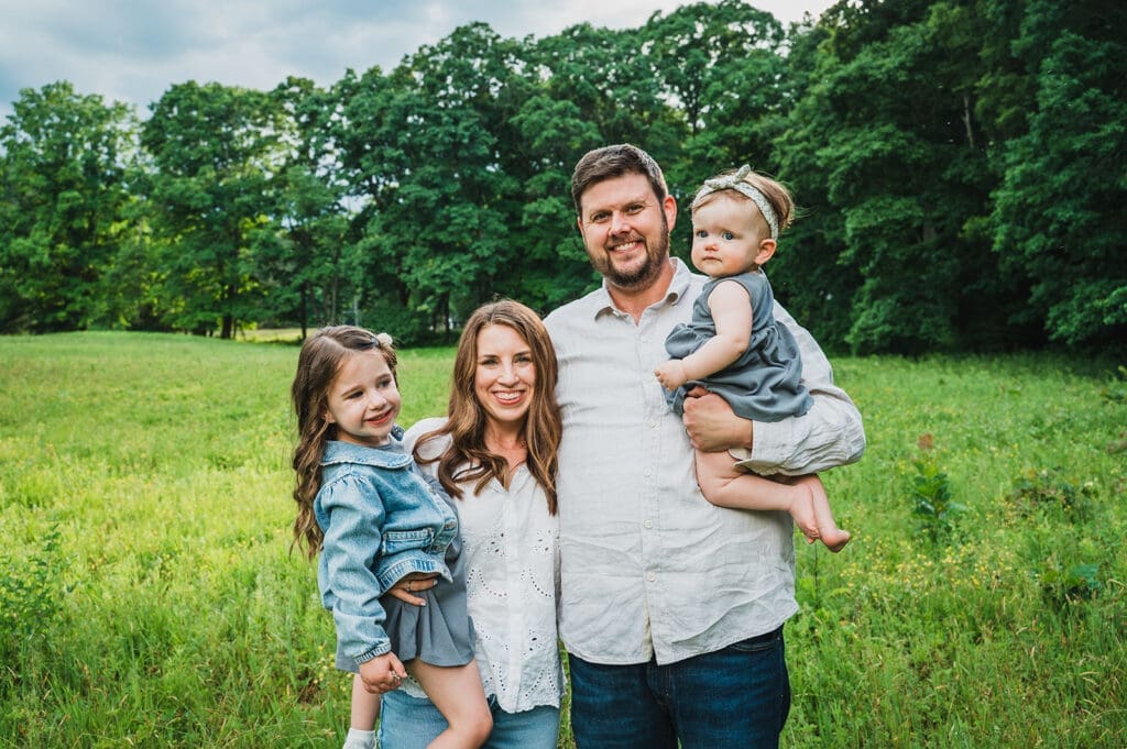 a family of four stands together in a sunny field smiling at the camera in well coordinated outfits of muted blue tones with creams and whites