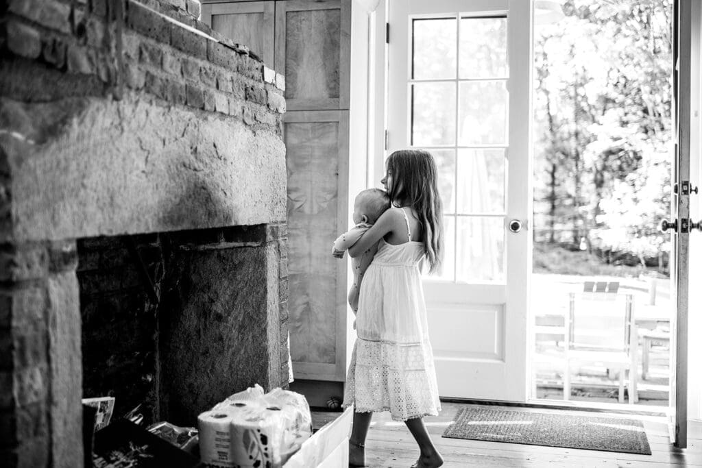 Girl carrying a baby at a doorway near a stone fireplace during a candid family photography session